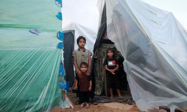 Three Palestinian children stand in between two make-shift tents of plastic