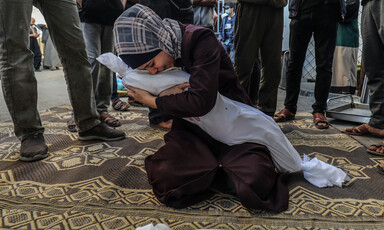 A woman kneeling on the ground cradles the sheet-wrapped corpse of a child