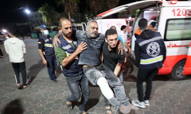 An injured man is carried by two others with an ambulance and medics in the background 