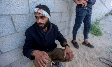 A man sit against a wall with a bandage covering his bleeding forehead while another man stands near him 