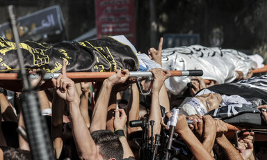 A view of hands carrying bodies wrapped in flags on stretchers while others hold rifles 