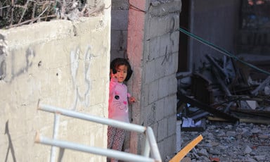 A child wearing a pink bunny sweater peeks out from behind a cinderblock wall surrounded by rubble