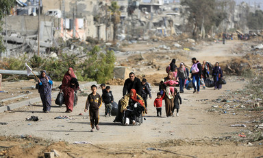 Landscape view of families, including woman carrying a white flag and a man pushing a woman in a wheelchair, walking on debris-strewn road with bombed-out buildings in the distance