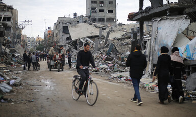 A man on a bicycle rides on a street interspersed with other people with bombed-out buildings on either side