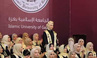 A young woman stands during a graduation ceremony in front of a banner featuring the logo of the Islamic University of Gaza 