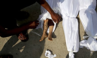 A person whose legs and arm are shown in the photo lifts a white sheet from the legs of a child's shrouded body