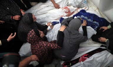Women sit next to and lean over several bodies shrouded in white sheets
