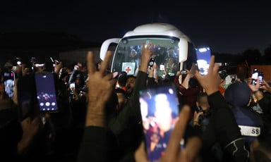 People hold their hands in the air, some making the V for victory gesture and others holding their phones, in front of a Red Crescent charter bus carrying freed women prisoners