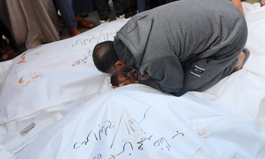 A man kneels down surrounded by dead bodies, most covered by white shrouds 