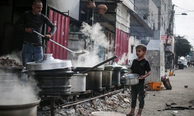A child waits with a bowl in front of pots of food cook over large wood-burning fires 