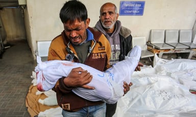 A man cries while he holds the shrouded body of a child as another man stands behind him and several shrouded bodies lay on the ground