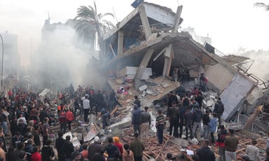 Dozens of people stand around bombed-out multi-story building