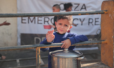 A boy with a pot in front of a banner with the words Gaza Emergency 