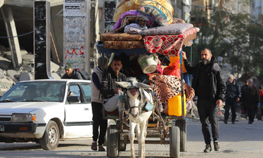 Two people walk alongside a donkey cart loaded with mattresses, jerry cans and other belongings