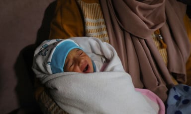 A baby wearing a blue cap is swaddled in a white blanket