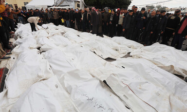 People line up at a funeral beside tens of bodies covered with white shrouds 