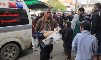 A man walks in front of an ambulance, carrying an injured toddler in his arms amidst a large crowd of people 