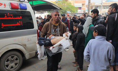 A man walks in front of an ambulance, carrying an injured toddler in his arms amidst a large crowd of people