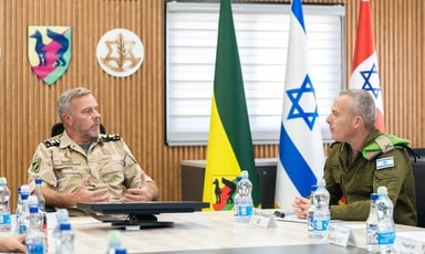 Two soldiers in uniform sit at desks and talk with three flags behind them