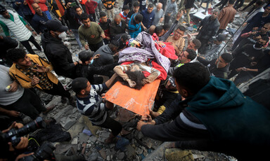 A child partly covered by a blanket is carried on a stretcher by several people amid a large crowd