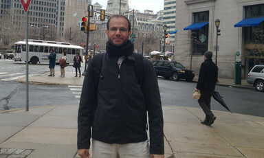 Refaat seen from waist up stands in front of a city crosswalk while wearing a black jacket and looking at the camera