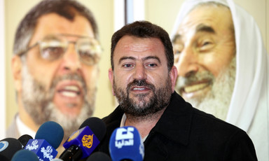 Saleh al-Arouri, seen from chest up, stands between news media microphones and banner showing Hamas leaders Abdel Aziz al-Rantisi and Sheikh Ahmed Yassin