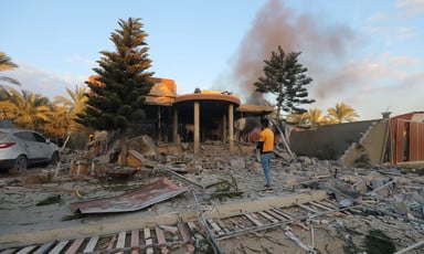 A man stands in front of a destroyed building, smoke rising from behind