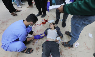 A man wearing medical scrubs holds a syringe above the arm of a boy laying on tiled floor smeared with blood