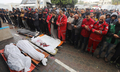 Mourners, some of them in red paramedic uniforms, pray over three bodies, wrapped in white shrouds. 