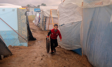 A boy carries a metal tray while walking on clay-like soil in between plastic-sheeted tents
