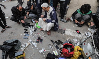 A group of men sit on the street around a power cord, charging phones