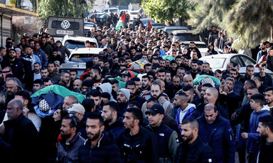 A large crowd of people carries shrouded bodies on stretchers in a street