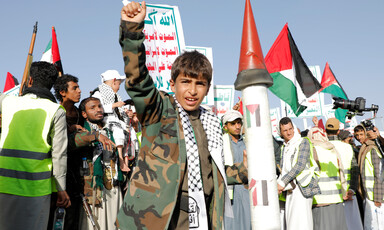 A boy wearing a camouflage jacket and a hatta holds his right fist in the air while standing in a crowd outside