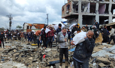 Men carry large sacks on their shoulders while walking on a flooded road covered with debris alongside bombed-out buildings