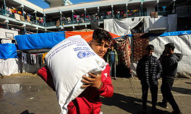 A Palestinian youth carries a bag of UNRWA flour with two youths and an UNRWA school in background