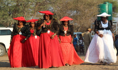 A group of women in bright flowing dresses and hats walk amid trees 