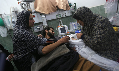 A woman with an injured face and a man laying on a hospital bed reach towards the shrouded body of baby held by a woman who is lifting fabric to expose the child's face