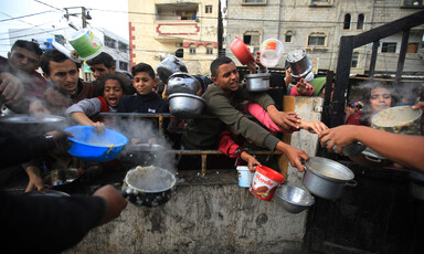 A crowd of men and children hold empty pots and buckets with outstretched arms