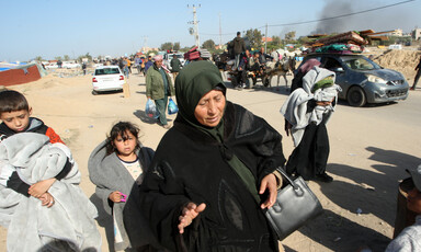 An older woman carries a small purse as she walks on sandy road with two young children carrying blankets close behind her and other people carrying their belongings traveling by foot, donkey cart and car at a distance behind her