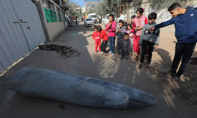 A group of young children in a street point at a large bomb lying on the ground