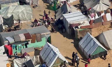Aerial view of people standing and sitting around improvised shelters covered with plastic on sandy ground