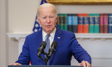 A man in a blue suit stands behind a podium with two microphones