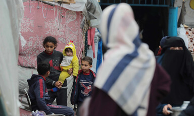 A group of children sit beside two women next to a makeshift tent in Gaza