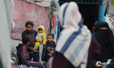 A group of children sit beside two women next to a makeshift tent in Gaza 