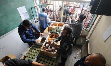 Men in a Gaza school stand beside boxes containing tinned foods as they prepare to hand them to people standing at the window