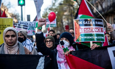 Woman wearing a protective face mask and kefffiyeh scarf raises a fist in the air while holding a Palestine flag amid crowd of protesters