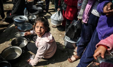 A girl sits on the ground with several empty tin pots. Other people are standing behind her in line