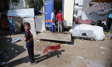 A young boy stands next to a blood stain on pavement and a truck being unloaded.