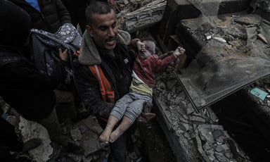 A civil defense worker holds the body of a toddler while standing on rubble