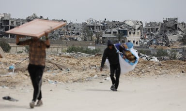 Two people carry some of their belongings on a road with the remains of homes that have been attacked in the background 